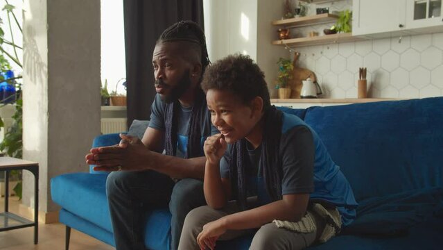 Excited adorable preadolescent African American boy and cheerful father in matching sports team shirts watching football match on tv, celebrating goal with winning gestures in domestic room.