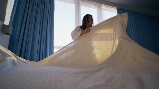 Woman in white bathrobe making morning routine, covers large bed with sheet after waking-up in spacious hotel room in sunny early morning. wide angle view