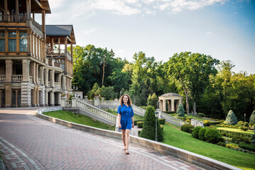 Beautiful young smiling woman walking in city park near famous old architecture building at summer day wearing dress and looking to the camera