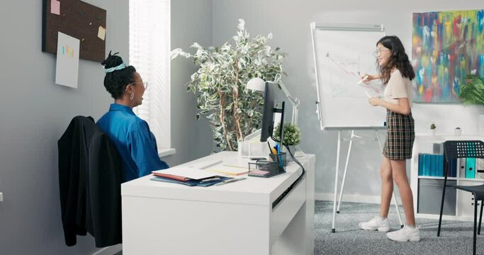 Two female in corporate office on break one of women is standing at chalkboard holding marker in hand, they agree to make joke and cross out lines, change appearance of drawing they laugh high-five