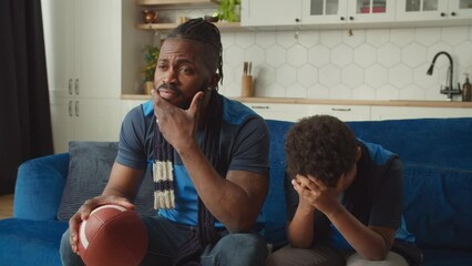 Emotional black father with American football ball and lovely school age boy in sports uniforms and scarves watching bowl game on tv, expressing disappointment and upset after team loss. - Powered by Adobe