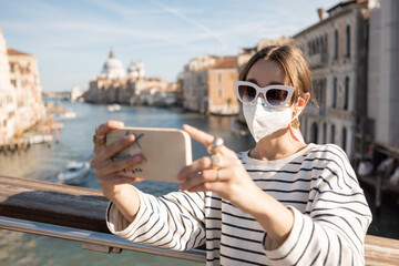Fototapeta na wymiar Young woman in medical mask taking selfie photo while traveling during pandemic in Italy. Concept of social rules of wearing a mask outdoors. Woman enjoying view on Grand canal in Venice