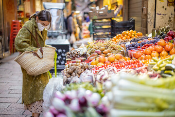Woman in medical mask buying fruits and vegetables at market stall on the famous gastronomical street in Bologna. Concept of buying local products during pandemic. Idea of Italian lifestyle