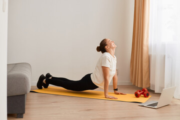 Indoor shot of fit woman making cobra pose on yoga mat, lying on her belly, places her palms under...