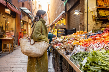 Woman in medical mask choosing fresh products at market stall on the famous gastronomical street in...