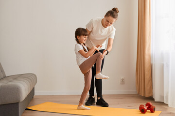 Fototapeta premium Indoor shot of of female sport trainer teaching little girl to do fitness exercises, kid standing on one leg and stretching, people posing in light room on mat.