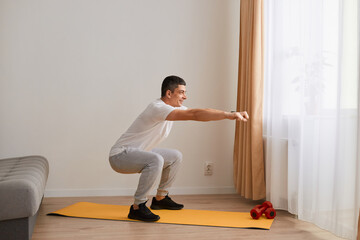 Side view portrait of athletic man wearing sportswear doing squat, lower body sport exercise, warming up and training muscles, keeps arms outstretched, looking ahead.