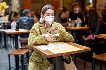 Woman in medical mask photographing qr code of restaurant menu while sitting on cafe terrace outdoors. Concept of Italian gastronomic culture and new social rules related to pandemic