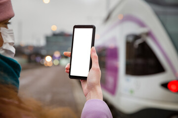 Smart phone with white screen in hand. Woman in protective mask showing phone display. Railway station platform with train. Person traveling and using phone app.