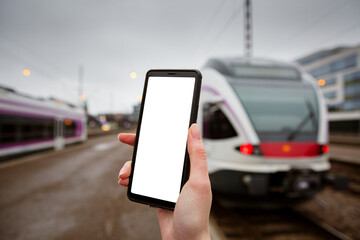 Close-up smart phone with white screen in hand. Railway station platform. Person traveling and using phone app. Trains on the background.
