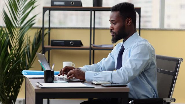 Young businessman or executive using laptop and writing, sitting at desk at home or office spbi. Close-up view of handsome African man types and looks at computer screen, writes in notebook and sits