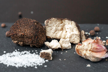 whole and broken ball of Belper Knoll cheese, black pepper, salt and garlic cloves lying on a black slate. Hard Swiss cheese in the form of small balls, sprinkled with black pepper, close-up.