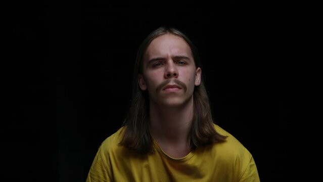 Portrait of a young guy with interest looking into the camera and showing a call me gesture. Male model with long hair and mustache posing on a black background in the studio. Close up. Slow motion.
