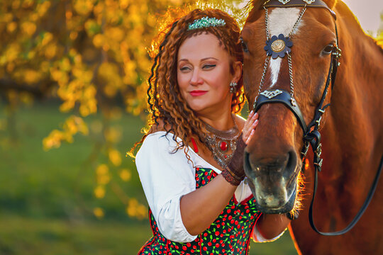 Close portrait of pretty woman with curly next to big brown horse in the autumn sunset light in Riga, Latvia. Woman  holding horse by the muzzle.