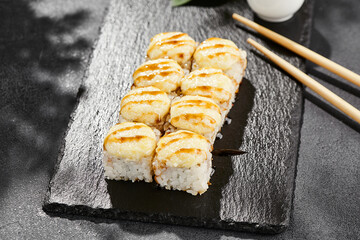 Baked Maki sushi on dark slate. Hot cheese maki with unagi sauce. Sushi roll with baked cheese and unagi sauce topped. Style concept japanese menu with black background, leaves and hard shadow.