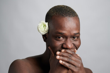 Young African man with white camelia flower by ear covering his mouth while laughing in front of...