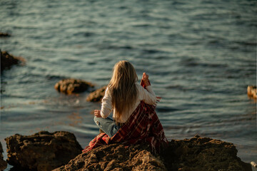 Attractive blonde Caucasian woman enjoying time on the beach at sunset, sitting in a blanket and looking to the side, with the sunset sky and sea in the background. Beach vacation.