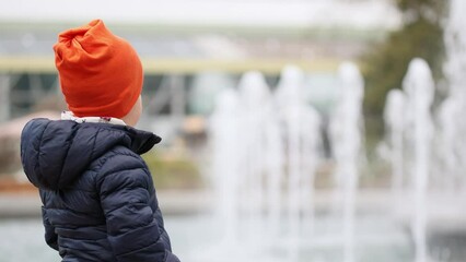 little caucasian boy orange hat blue coat looking working water fountain in cold sunny day city park. child kid admiring fountain spray splash looking camera hands in pocket. autumn spring weather 