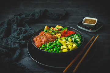 Japanese poke or buddha bowl with salmon, avocado, roe, mango, pies and cucumber in a black bowl on black background