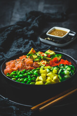 Japanese poke or buddha bowl with salmon, avocado, roe, mango, pies and cucumber in a black bowl on black background, vertical