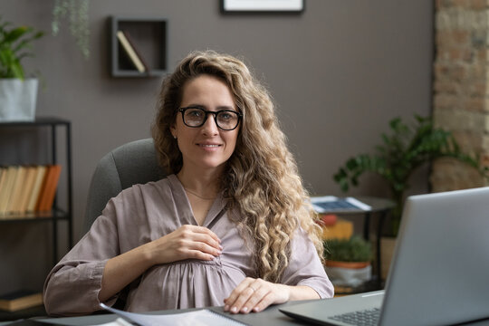 Happy young pregnant businesswoman in casualwear sitting by desk in frot of laptop and looking at camera with smile