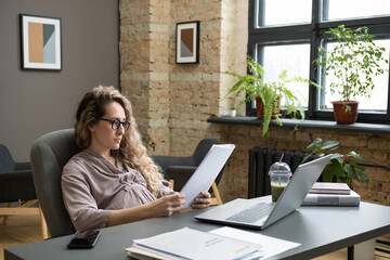 Young pregnant businesswoman looking through financial papers in front of laptop while sitting by workplace in office