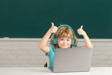 Clever school boy with thumbs up, cute pupil wears headphones writing on laptop, listen audio lesson use computer. Videocall, e-study with tutor distantly. First day at school.