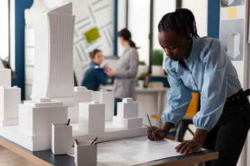 Residential architect drawing blueprints at desk in architectural office next to white foam scale model of skyscraper. Development engineer making notes on construction plan.