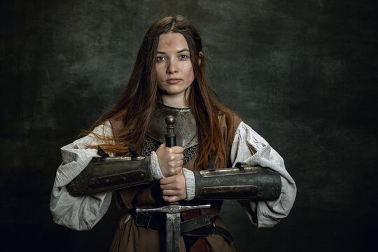 Vintage portrait of adorable woman, medieval female warrior or knight with dirty wounded face looking at camera isolated over dark retro background.