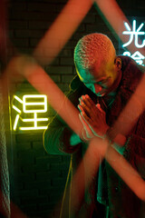 Obraz premium Young African man in furcoat keeping his hands put together and bowing while standing behind bars against illuminated hieroglyphs