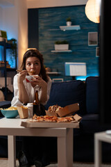 Woman sitting on sofa watching television while eating takeout asian noodles with chopsticks in home living room. Office worker enjoying delicious tv dinner fast food delivery box.