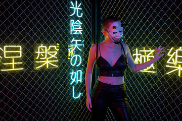 Young cyberpunk female in monster mask and black leather clothes holding by bars while standing in...