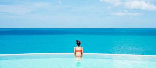 Happy woman in orange swimsuit swimming in infinity pool at luxury hotel against ocean front. young...