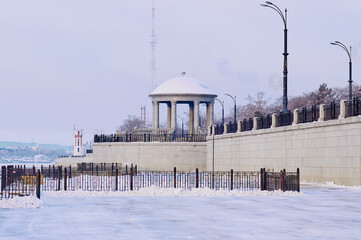 River embankment in snowy winter. The snow-white dome of the rotunda with marble columns and a balustrade. White lighthouse and TV tower. Metal fences and lanterns on the promenade