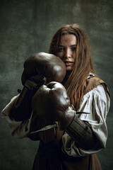 Portrait of brave girl, medieval warrior or knight with dirty wounded face in boxing gloves isolated over dark vintage background. Comparison of eras, history