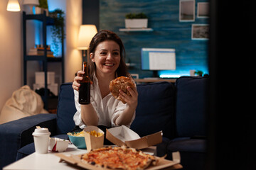 Woman laughing watching television holding lager beer bottle and takeaway fast food tasty hamburger sitting on couch in livingroom. Smiling happy person having takeout order pizza and junk food.