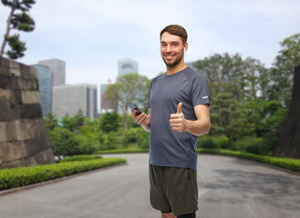 Fototapeta premium fitness, sport and healthy lifestyle concept - smiling man in sports clothes with smartphone and earphones listening to music and showing thumbs up gesture over park in tokyo city, japan on background
