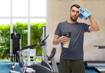 fitness, sport and healthy lifestyle concept - tired man in sports clothes with protein shake bottle wiping his forehead with towel over gym background