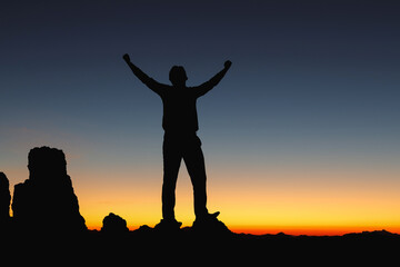 Fototapeta na wymiar Successful people silhouette with open arms on rock morning sunset sky background.