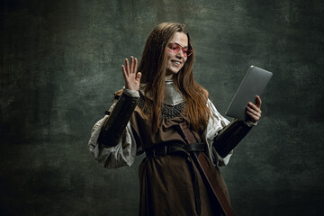 Vintage portrait of young beautiful girl in image of medieval warlike woman using modern gadget isolated over dark background. Comparison of eras, history