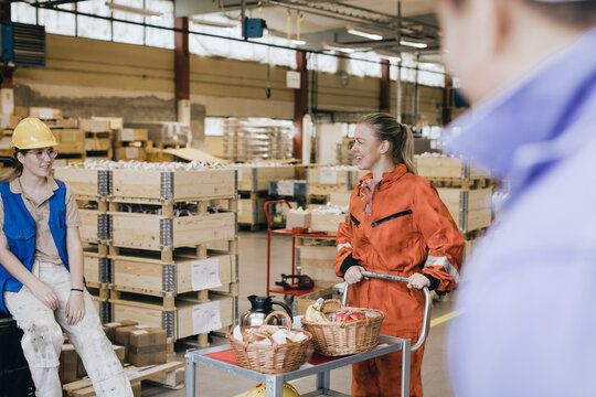 Smiling young female worker with food on push cart talking with colleague in warehouse