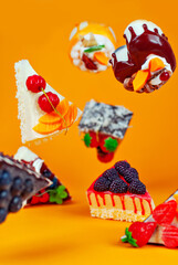 Various cakes fall on an orange background. Glazed triangular pieces of cake with chocolate filling and fruit creams. Sweets fly in the air.