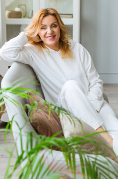 Indoor portrait of attractive European woman in her 40plus with blonde hair and fit body looking at camera with pleased friendly smile, sitting on comfortable gray couch
