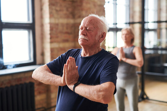 Senior man standing in yoga pose with his eyes closed and meditating during class with other people in health club