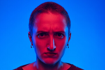 Close-up angry girl, student with short hair looking at camera isolated on dark blue background in neon light. Concept of emotions, gender x