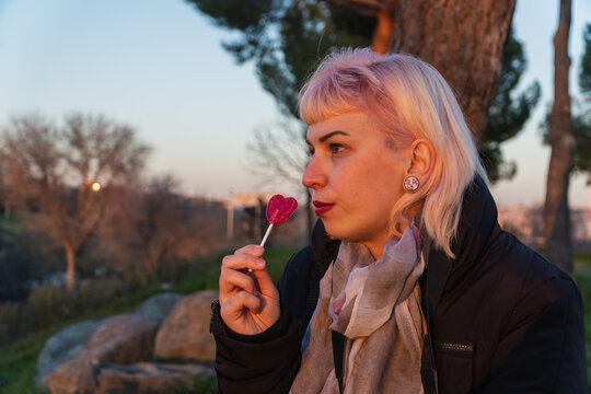 El beneficio de la glucosa es despertar nuestra actividad, Chica sentada en un parque comiendo una piruleta, chica con cabello rosa sentada en un parque mirando al horizonte mientras come una piruleta