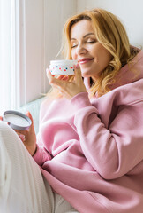 A mature woman in a pink hoodie sits on the windowsill, holds a jar of cream in her hands and sniffs it.