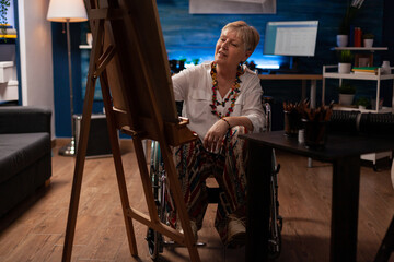 Elderly woman sitting in wheelchair drawing black and white sketch using pencil on canvas in artist studio. Senior citizen who is paralyzed feeling creative in front of easel creating fine art.