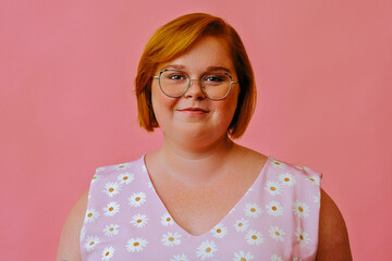 headshot of smiling young redhead woman with short hair and eyeglasses looking at camera in studio over pink background - obrazy, fototapety, plakaty