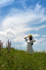 Brunette in dress stands in middle of green meadow and holds wreath of wildflowers on her head with her hands, enjoying moment with her eyes closed. Free life. Selective focus.